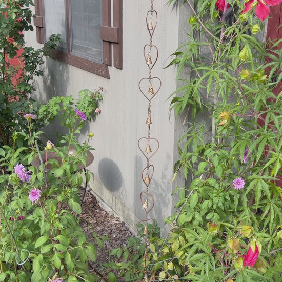 A decorative metal rain heart rain chain with interlocking heart shapes hangs from the eaves of a white porch with columns and a dark-framed front door.