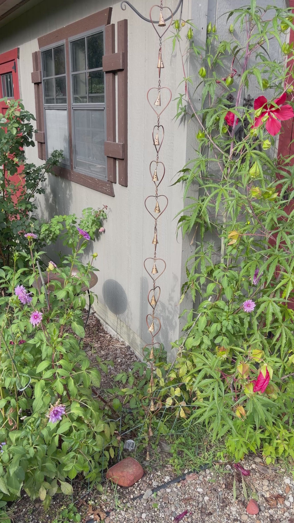 A decorative metal rain heart rain chain with interlocking heart shapes hangs from the eaves of a white porch with columns and a dark-framed front door.