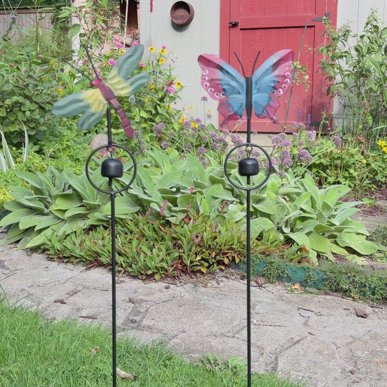 A butterfly-shaped metal garden stake with a hanging bell, featuring red and blue patterned wings, photographed against a gray wooden fence.