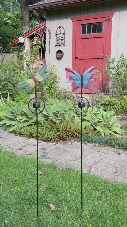 A butterfly-shaped metal garden stake with a hanging bell, featuring red and blue patterned wings, photographed against a gray wooden fence.