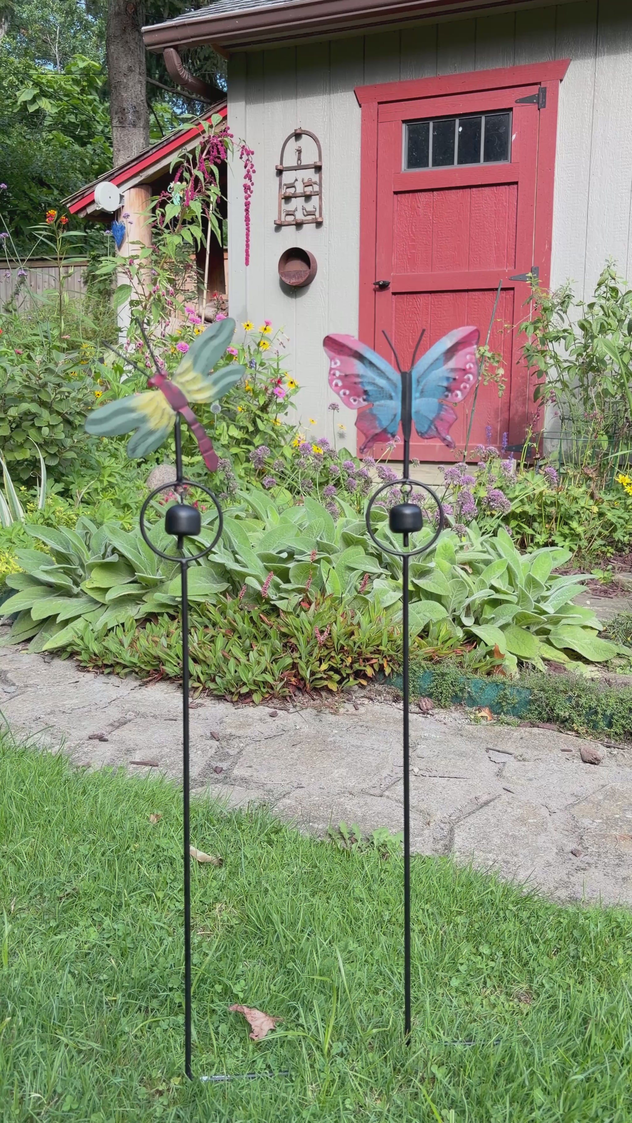 A butterfly-shaped metal garden stake with a hanging bell, featuring red and blue patterned wings, photographed against a gray wooden fence.