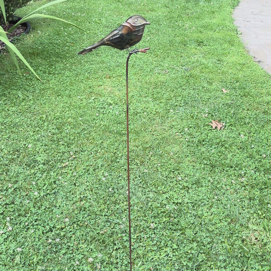 Copper-toned metal bird sculpture perched on a thin rust-colored stake, set against green foliage