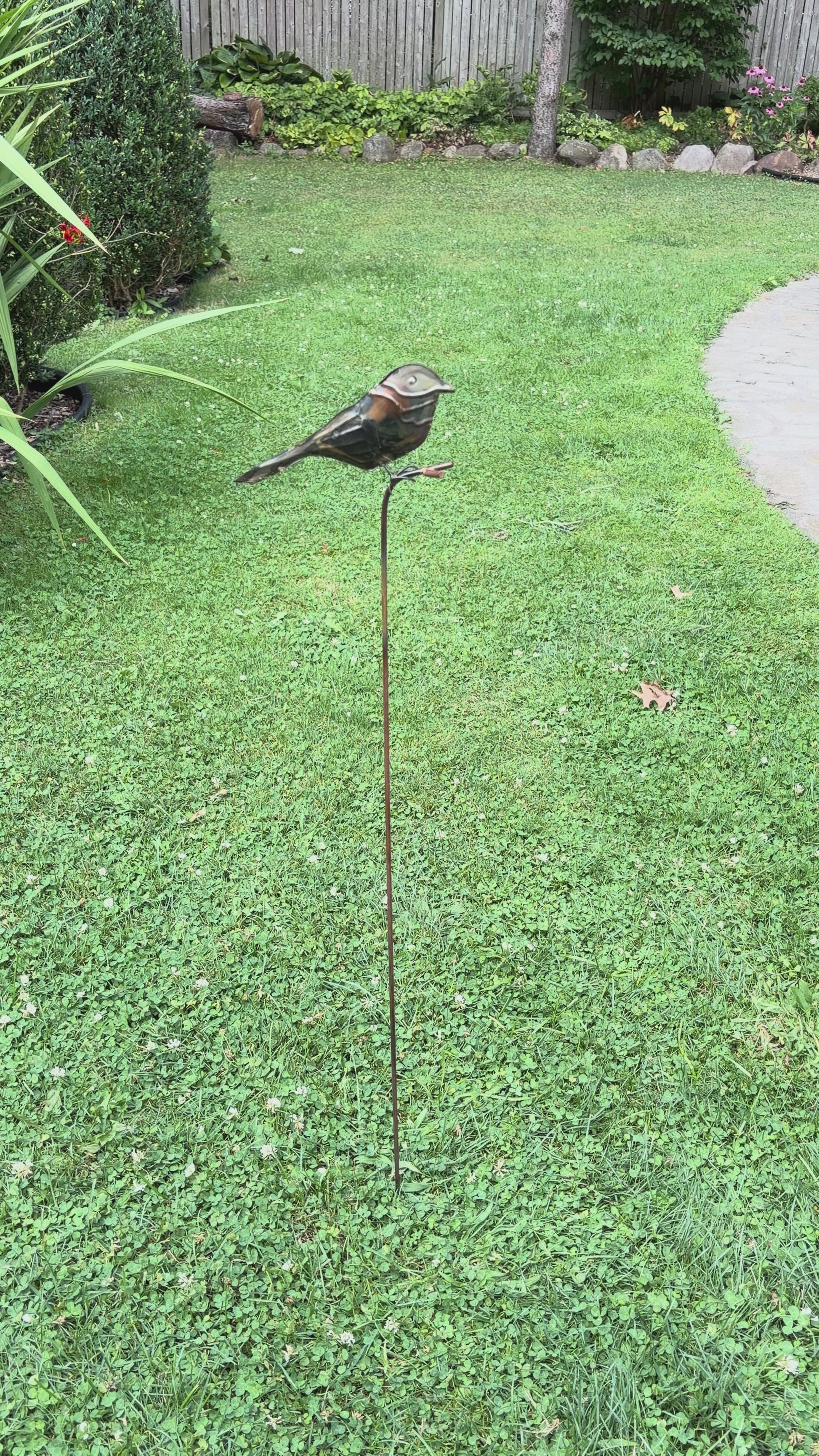 Copper-toned metal bird sculpture perched on a thin rust-colored stake, set against green foliage