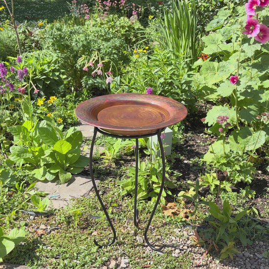 Hammered copper round birdbath with iridescent finish on curved metal stand.