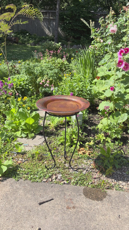 Hammered copper round birdbath with iridescent finish on curved metal stand.