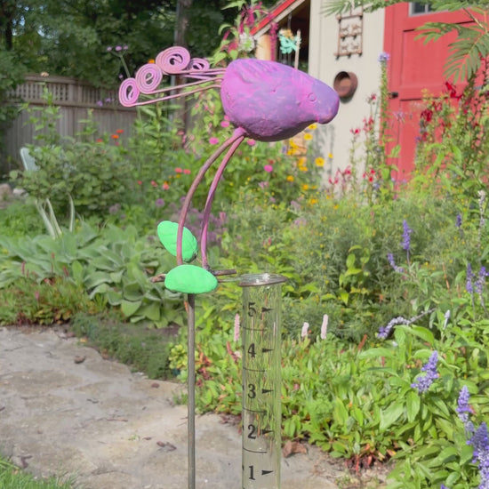 A decorative metal garden stake shaped like a purple bird with green leaves and curled purple stems, mounted on a clear glass test tube, photographed outdoors on grass beside a stone path.
