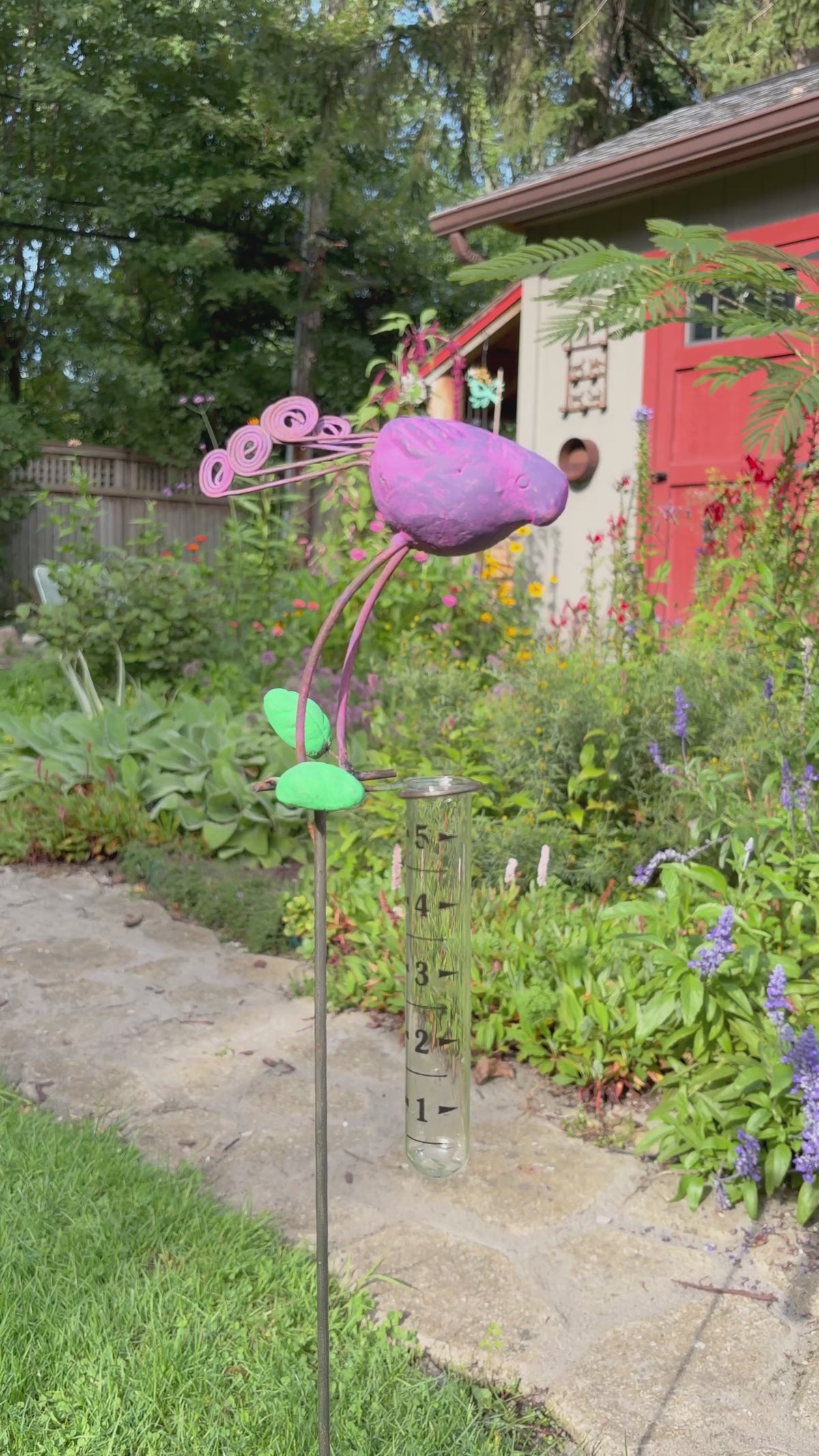 A decorative metal garden stake shaped like a purple bird with green leaves and curled purple stems, mounted on a clear glass test tube, photographed outdoors on grass beside a stone path.