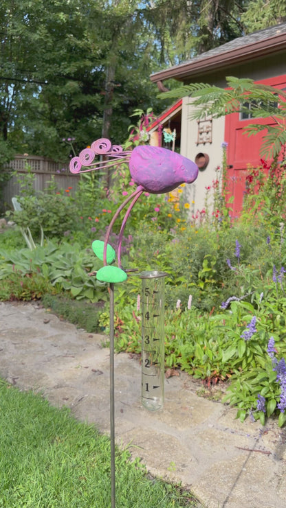 A decorative metal garden stake shaped like a purple bird with green leaves and curled purple stems, mounted on a clear glass test tube, photographed outdoors on grass beside a stone path.