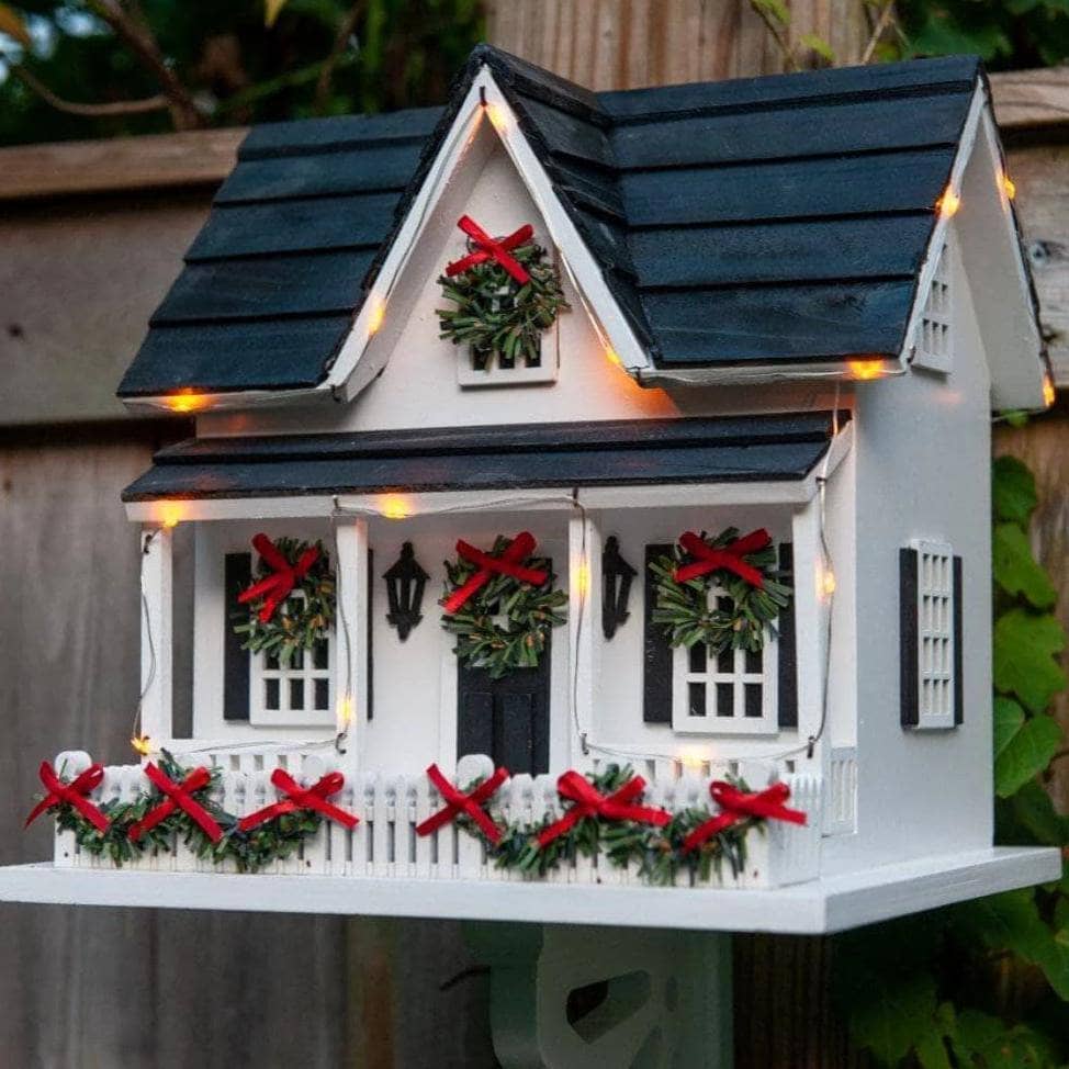 White miniature cottage birdhouse with dark roof, LED string lights, mini wreaths with red bows, and white picket fence
