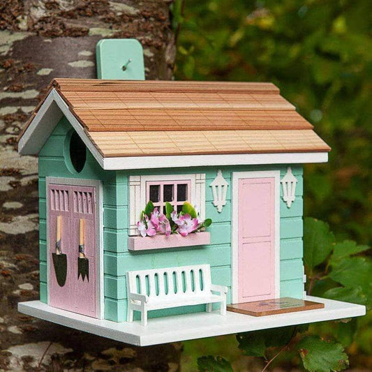 Mint green wooden birdhouse shaped like a cottage with pink doors, flower window box, white bench, and cedar-style roof