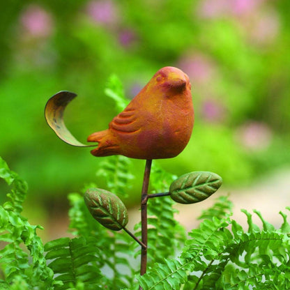 Rusty orange terracotta bird figurine on a metal stake with green cast leaf accents, placed among fern plants