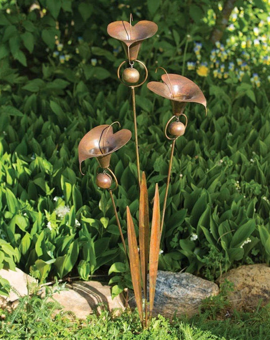 Copper-toned metal calla lily flower garden stake with a bell and curled wire detail, placed among green leafy plants