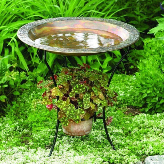 Copper-finish round bird bath bowl on black wrought iron stand, placed in a mulched garden bed with purple flowers